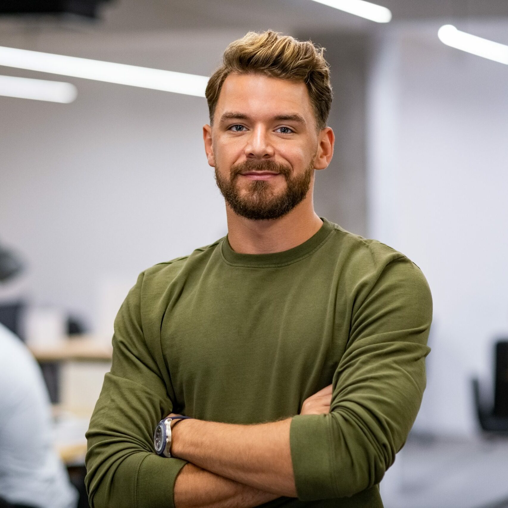 Portrait of proud businessman standing in creative modern office and looking at camera. Successful mid man entrepreneur standing with staff working in background. Satisfied young business man with crossed arms in advertising agency.